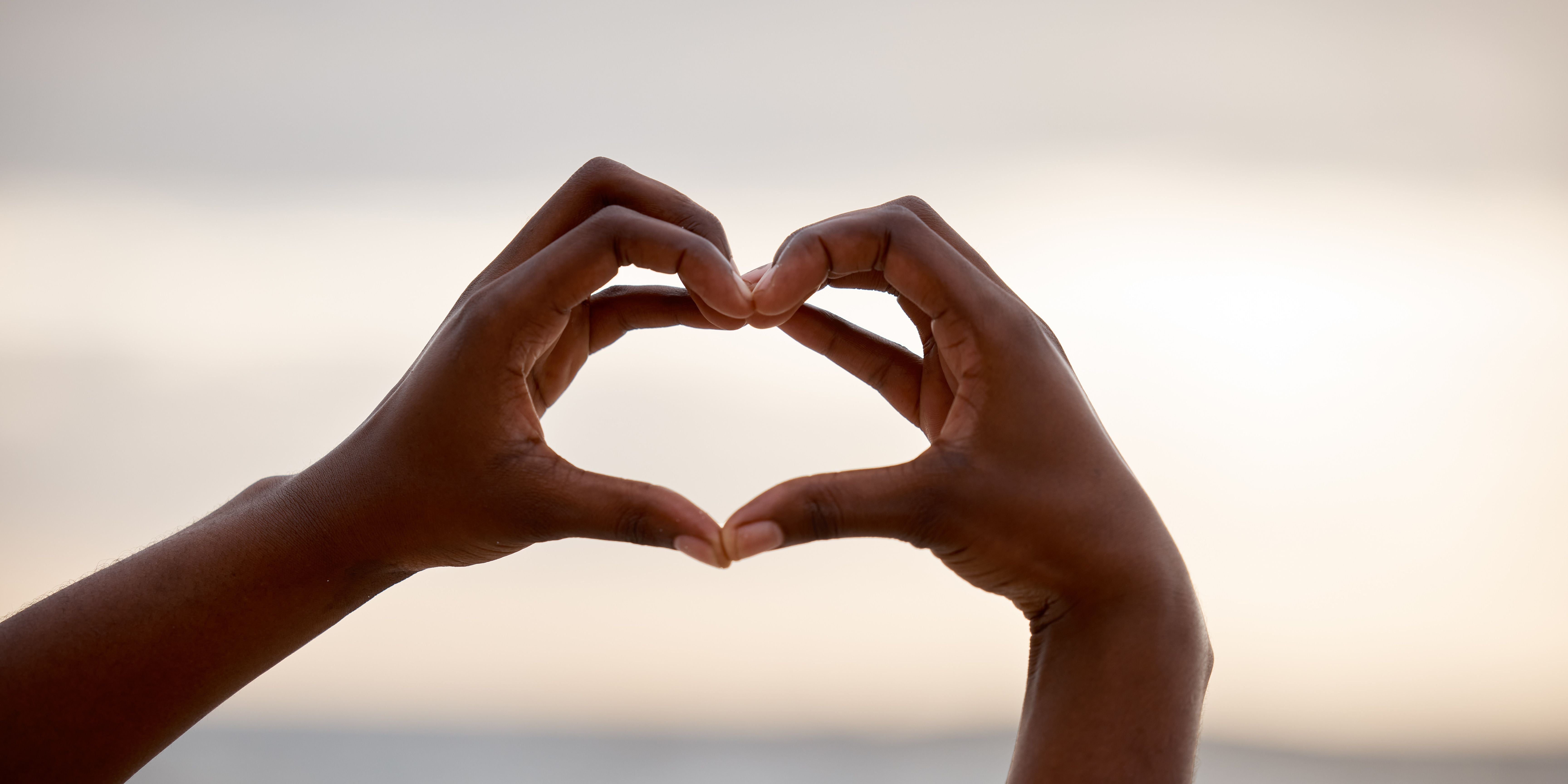Image of hands connecting and creating a love heart shape against a seashore skyline 