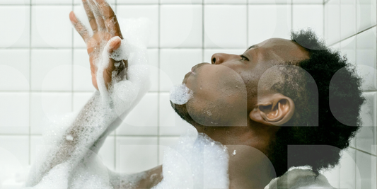 A young man reclining in a bubble bath, head tilted back with eyes closed, foam-covered hand raised against white tiles