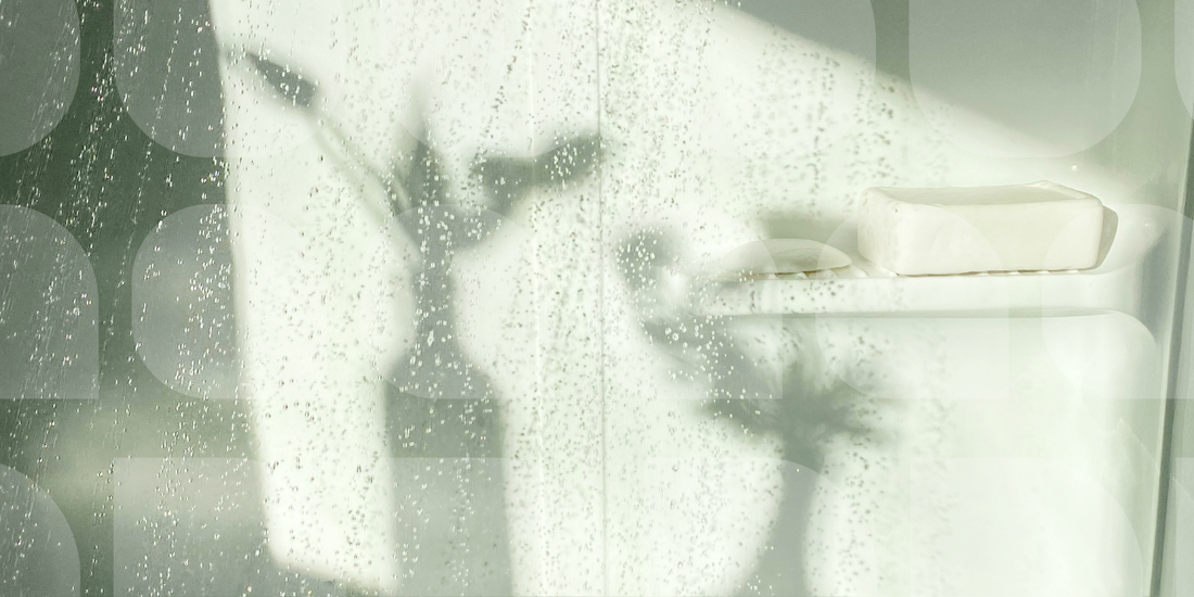 Close-up of a water-spotted glass shower door with a white soap bar on the ledge and soft plant shadows visible through the steam