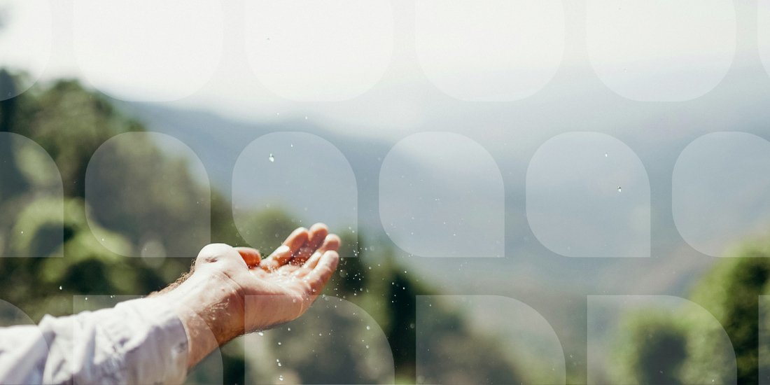 An outstretched hand catching water droplets outdoors against a soft-focus green landscape and misty mountain backdrop
