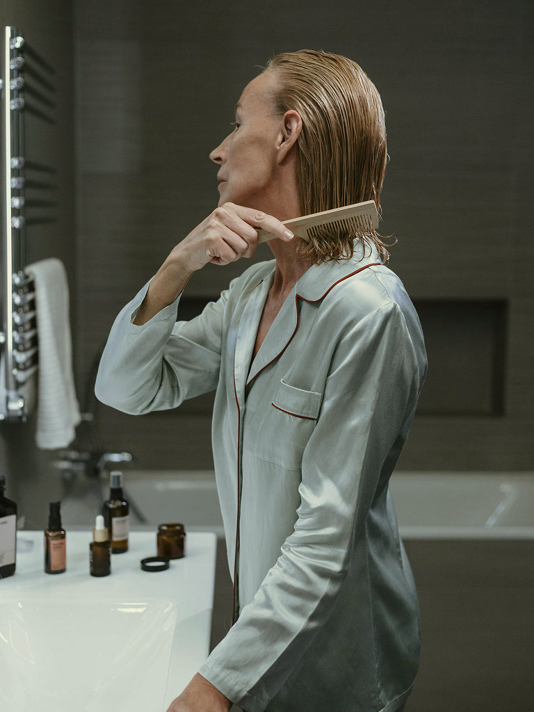 Woman brushing her wet hair in a bathroom with various bottles on a counter.