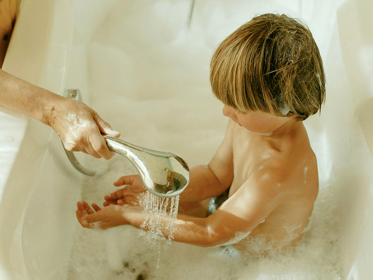 Child being bathed by an adult with a handheld shower head in a bathtub.
