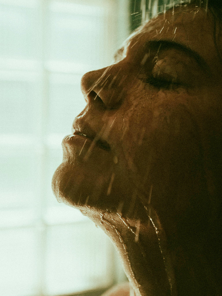 Close-up of a person's face with shower water pouring on their skin, looking out a window.