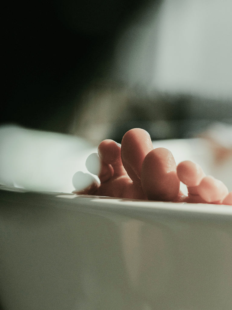 Close-up of toes resting on the edge of a white bathtub with soap bubbles, against a soft dark background