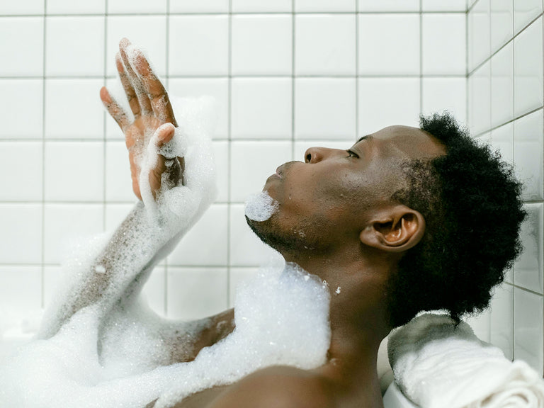 A young man relaxing in a bubble bath, leaning back with eyes closed, hand raised blowing the bubbles off