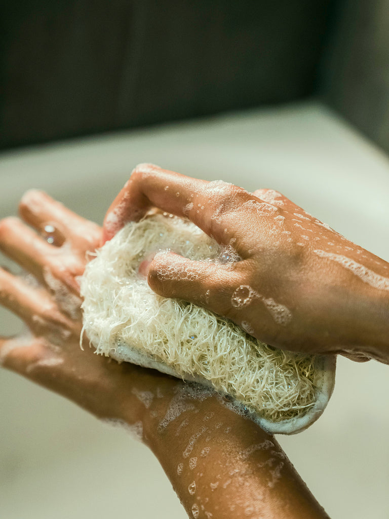 womans hand holding a loofah sponge with soap suds and water droplets on a neutral background