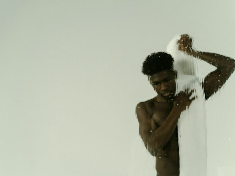 Man visible through a wet shower screen with a white towel draped over his shoulder against a plain background